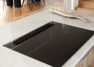 Black induction cooktop with a glass splash guard set into a stone benchtop with timber cabinetry, overlooking a dining area with a black table and chairs